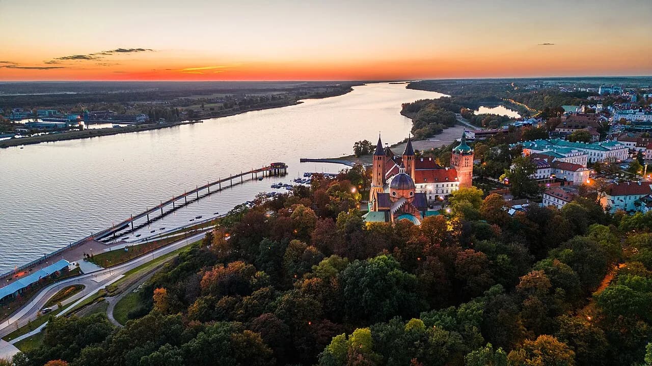 An aerial view of a town built along a river at sunset, featuring a large cathedral with multiple spires and a historic pier.