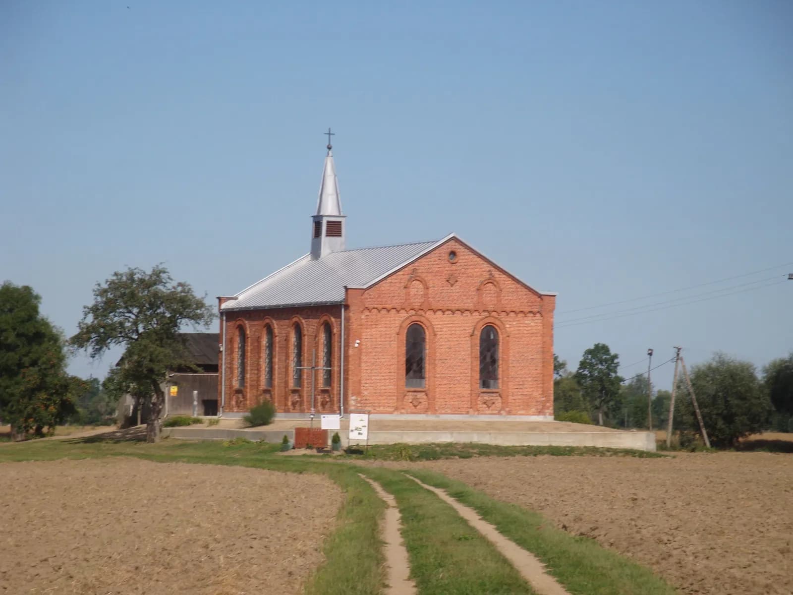 A brick church with a silver steeple and roof stands in a field under a clear blue sky, with a dirt path leading towards it.